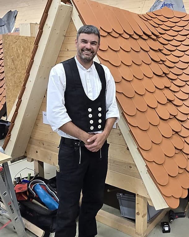 Dachdeckermeister des Betriebs in Zunftkleidung vor einem Dachmodell mit Biberschwanzdeckung
